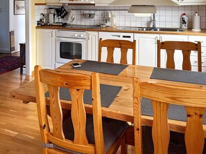 Uma sala de jantar com uma mesa e cadeiras de madeira, uma cozinha ao fundo e um tapete vermelho no chão.