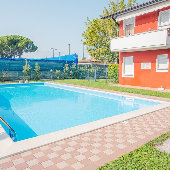 A swimming pool with a tiled deck, a red building with white trim, and a blue fence in the background. - Sweet Garden Apartment - Italian Homing (Sirmione)
