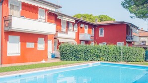 A swimming pool with a clear blue water surface, surrounded by a red building with balconies and a hedge in the foreground.