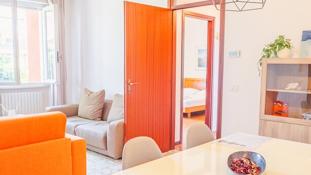 A modern hotel room with a white bed, orange sofa, and a dining table with a bowl of fruit.