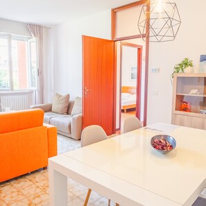 A modern hotel room with a white bed, orange sofa, and a dining table with a bowl of fruit. - Sweet Garden Apartment - Italian Homing (Sirmione)
