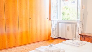 A bedroom with a wooden wardrobe, a bed with white linens, a radiator, and a window with curtains.