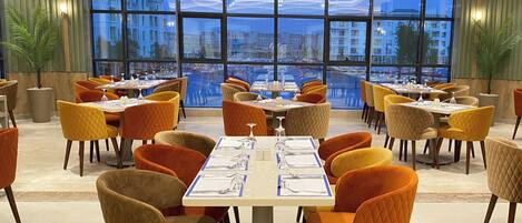 A dining area with tables and chairs, large windows with a city view, and potted plants.