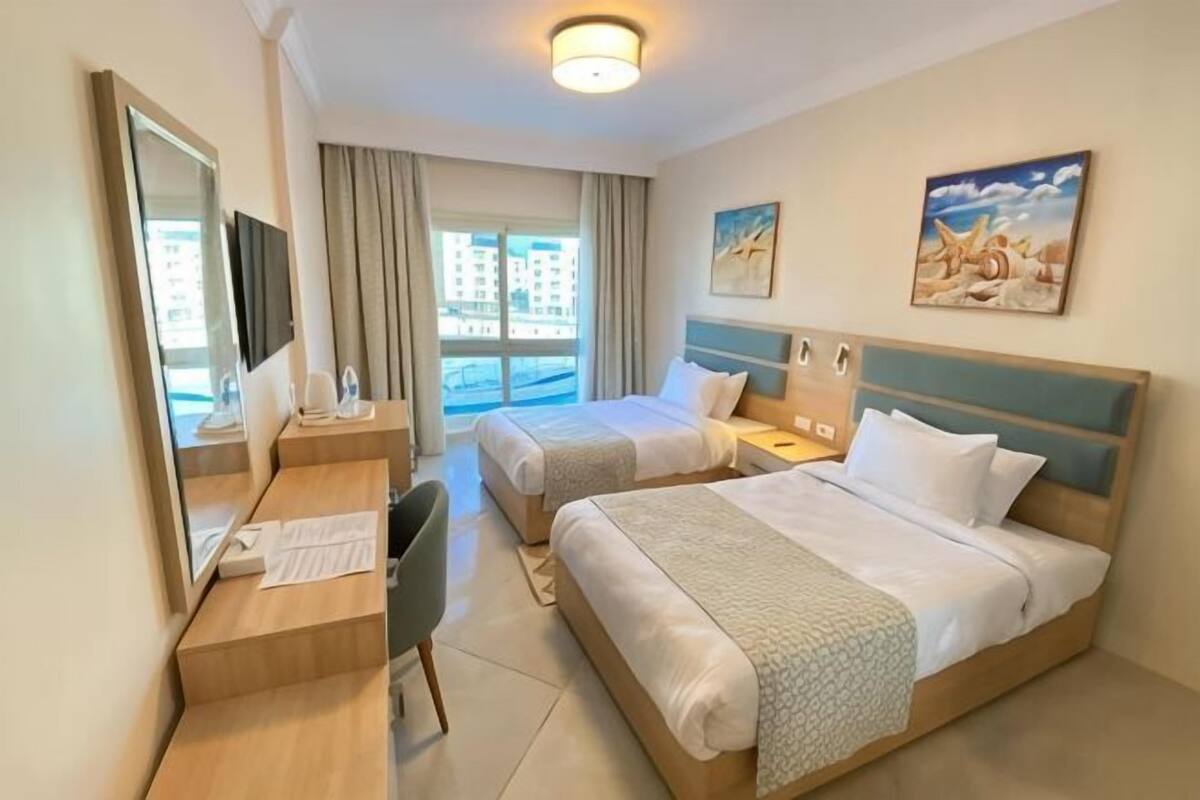 A hotel room with two beds, a desk, a chair, a TV, and a window with curtains.