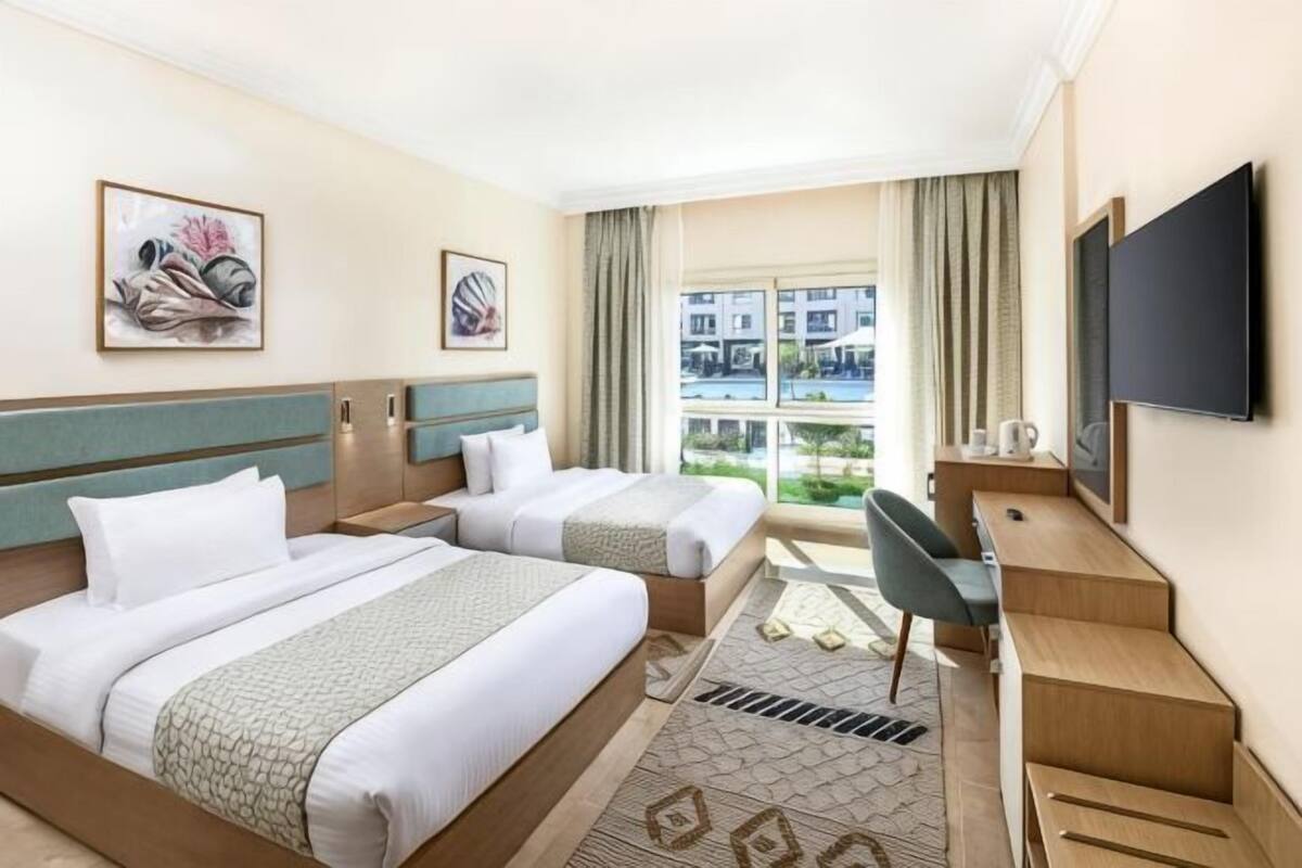 A hotel room with two beds, a desk, a TV, and a window with a view of buildings.