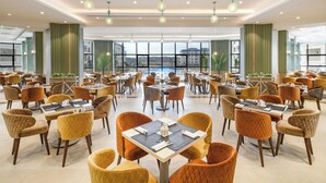 A spacious dining area with multiple tables and chairs, large windows offering a view of the city, and modern lighting fixtures. - Azur One Eleven - New Alamein (El Alamein)