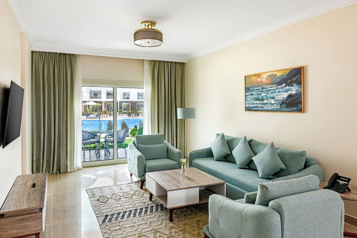 A living room with a sofa, armchairs, a coffee table, a flat-screen TV, a painting, and a view of a pool and outdoor area.