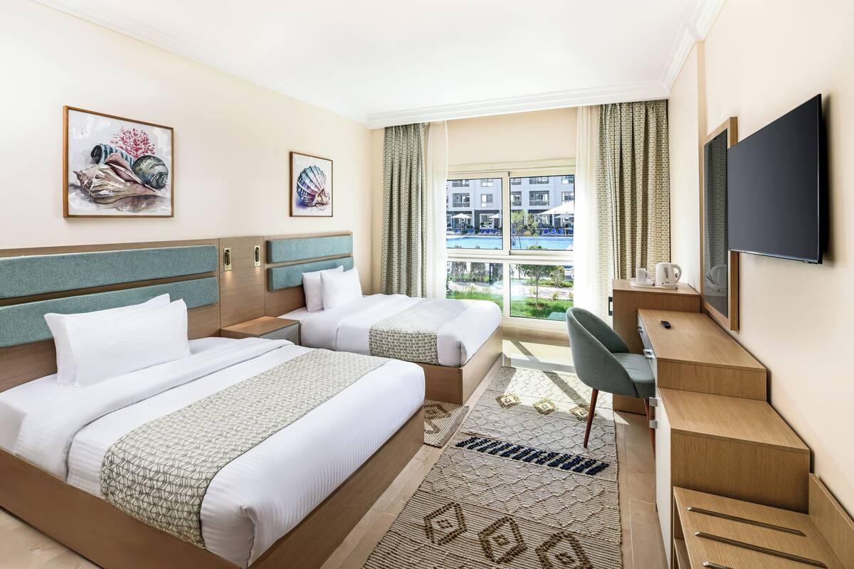 A hotel room with two beds, a desk, a TV, and a view of a swimming pool.