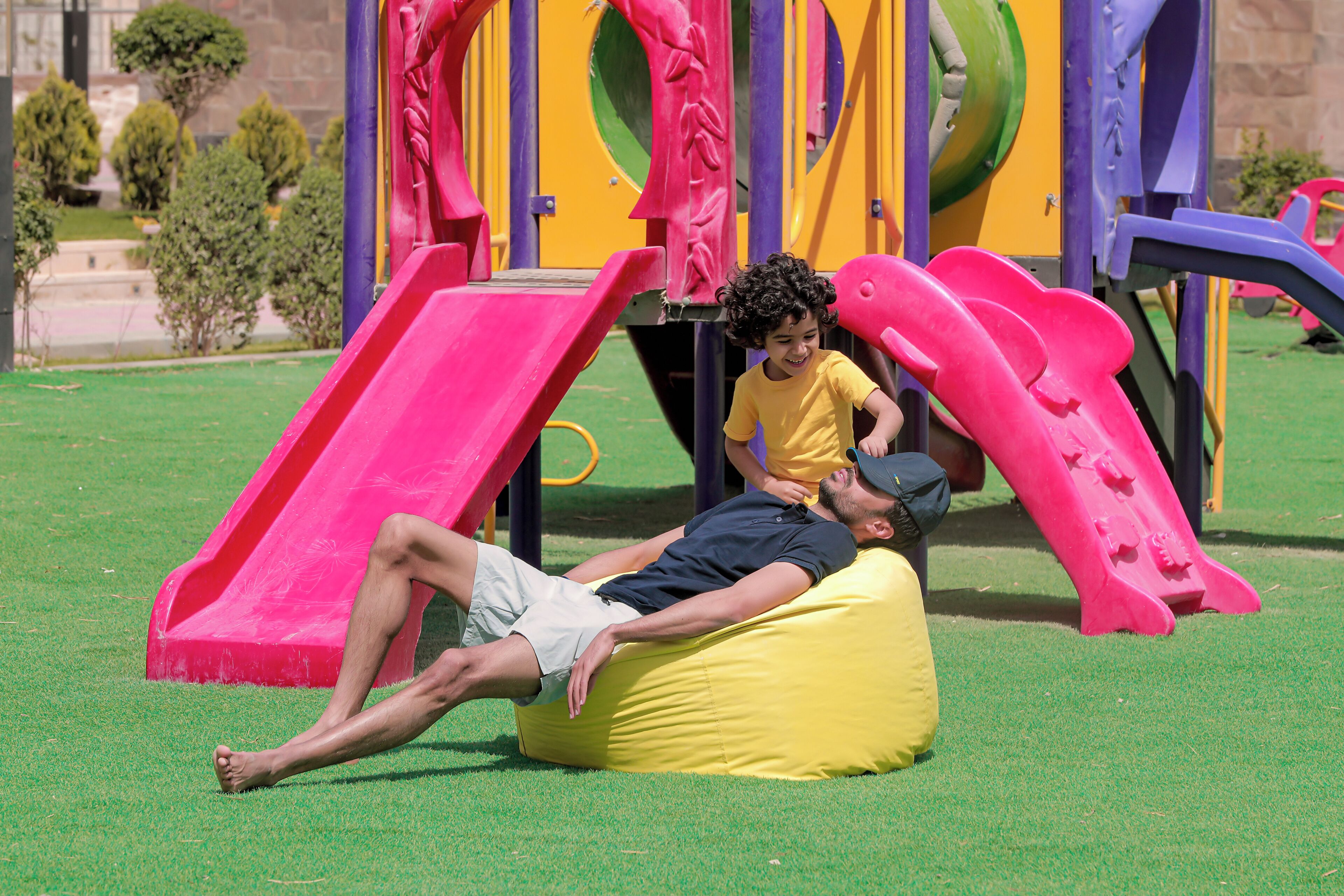 Un hombre recostado en un sillón de mimbre amarillo sobre césped, con un parque infantil al fondo.