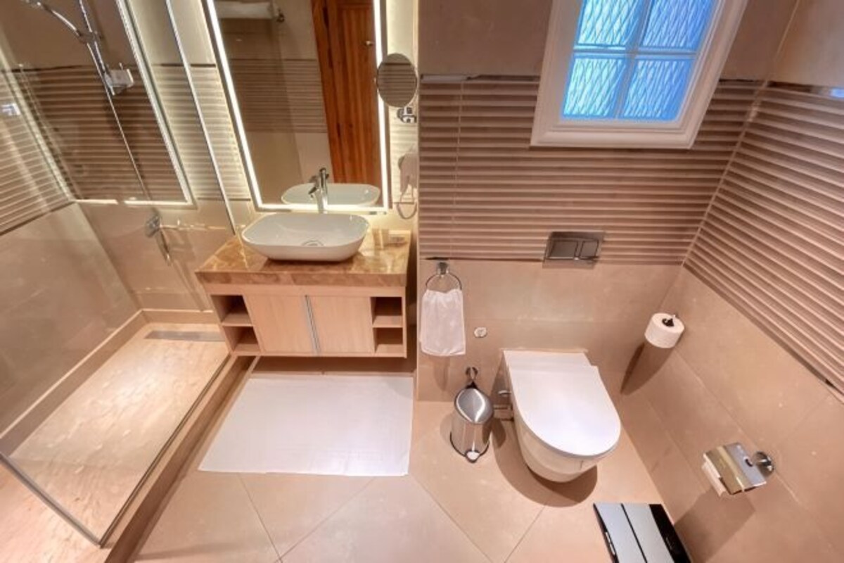 Un baño moderno con cabina de ducha de vidrio, un lavamanos con espejo, un inodoro y una ventana que deja entrar luz natural.