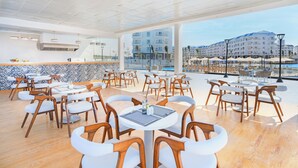 A modern outdoor dining area with wooden chairs and tables, a tiled backsplash, and a view of a pool and buildings. - Azur One Eleven - New Alamein (El Alamein)