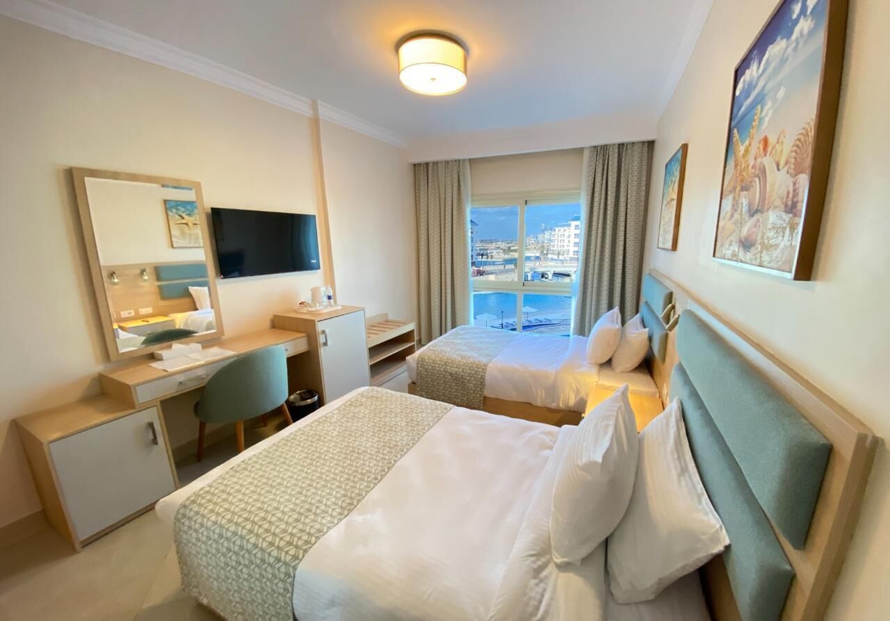 Ein Hotelzimmer mit zwei Betten, einem Schreibtisch, einem Stuhl, einem Fernseher und einem großen Fenster mit Blick auf die Stadt.