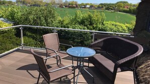 A deck with wicker furniture and a glass table, overlooking a green landscape with houses. - Farsighted Modern Retreat (Taarstedt)