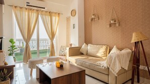 Ein Wohnzimmer mit einem beigefarbenen Sofa, einem Holztisch und einer Weidenlampe. An der Ziegelwand befindet sich ein Wandregal mit einer Pflanze und eine Uhr.
