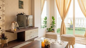 Ein modernes Wohnzimmer mit Flachbildfernseher, Couchtisch aus Holz und zwei Korbo ttomanen. Blumentöpfe und ein großes Fenster mit Vorhängen sind ebenfalls vorhanden.