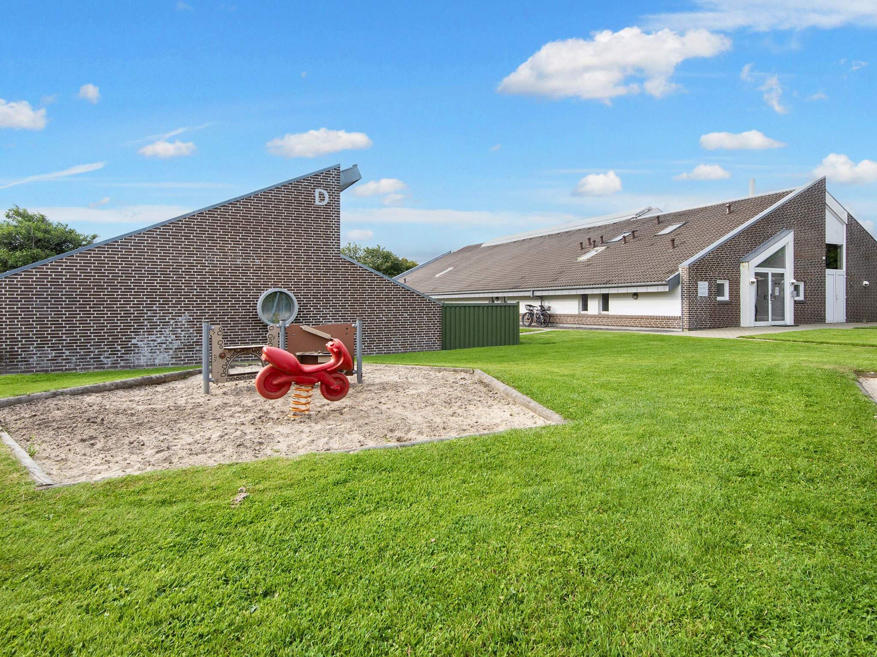 Une aire de jeux avec un scooter rouge et un bac à sable, un mur de briques et un bâtiment doté d’un toit en pente.