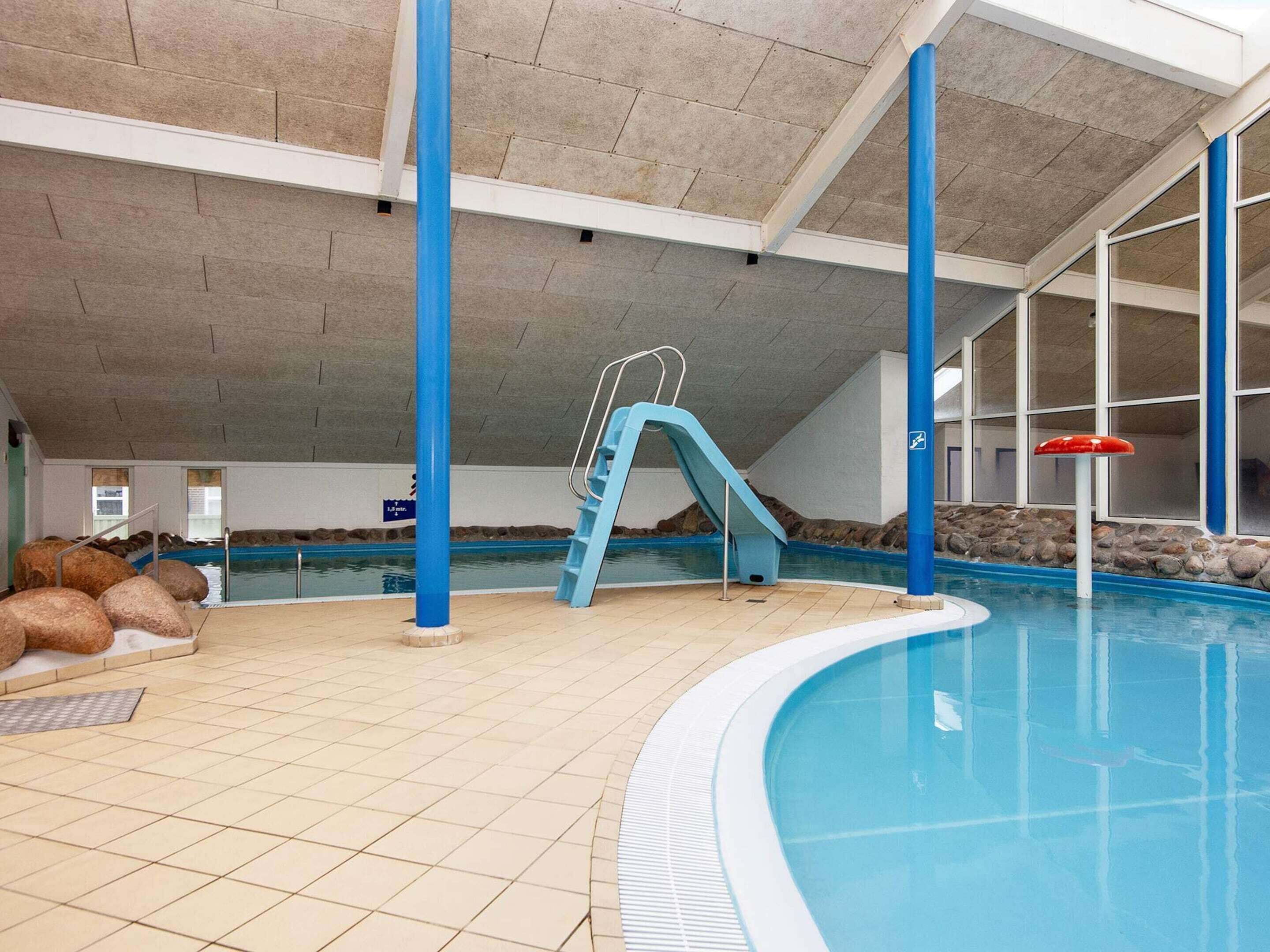 An indoor swimming pool with a blue slide, tiled floor, and large windows.