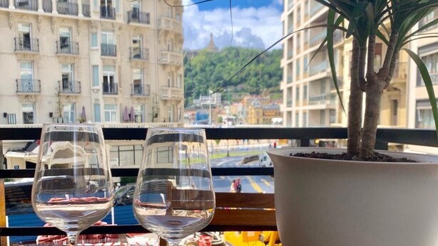 A balcony with two empty wine glasses, a potted plant, and a view of buildings and a road. - NEW Gorgeous Central Apartment - Beachfront - 3 BD (SAN SEBASTIAN)