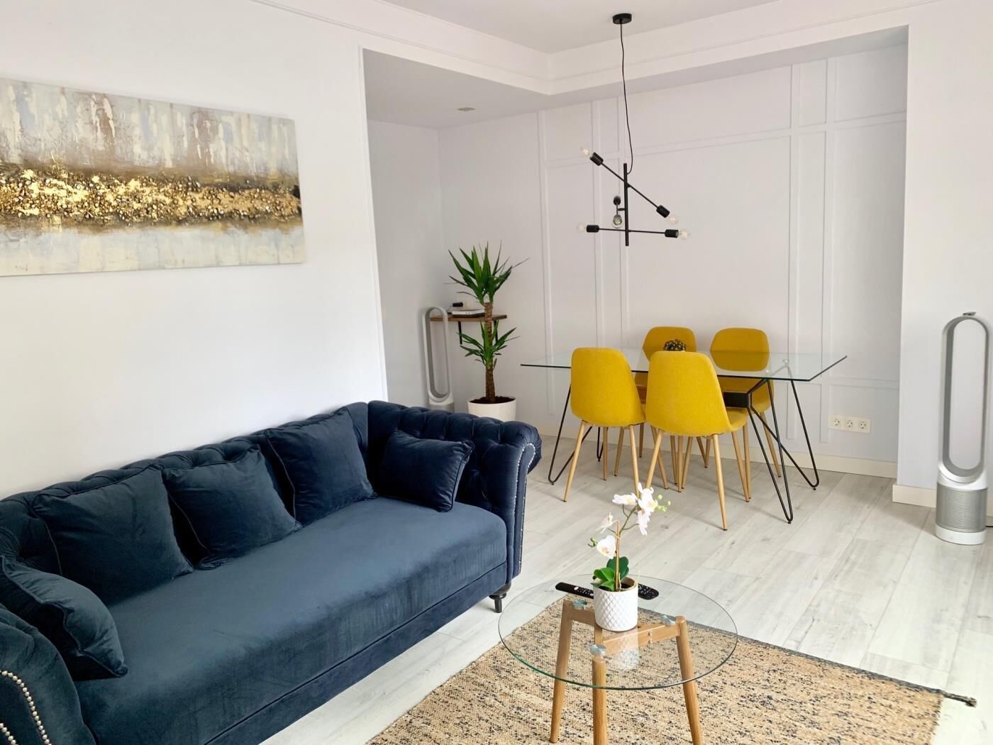 Un salon moderne avec un canapé bleu foncé, une table basse en verre et un coin repas avec des chaises jaunes.