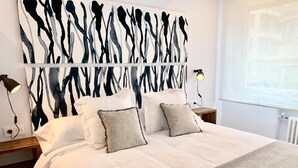 A modern bedroom with a large abstract painting on the wall, a bed with white linens and two pillows, bedside lamps, and a window with blinds. - NEW Gorgeous Central Apartment - Beachfront - 3 BD (SAN SEBASTIAN)