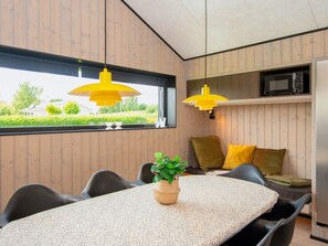 A modern kitchen with wooden paneling, a dining table with chairs, a yellow pendant light, and a view of greenery outside. - 8 Person Holiday Home in Hejls (Hejls)