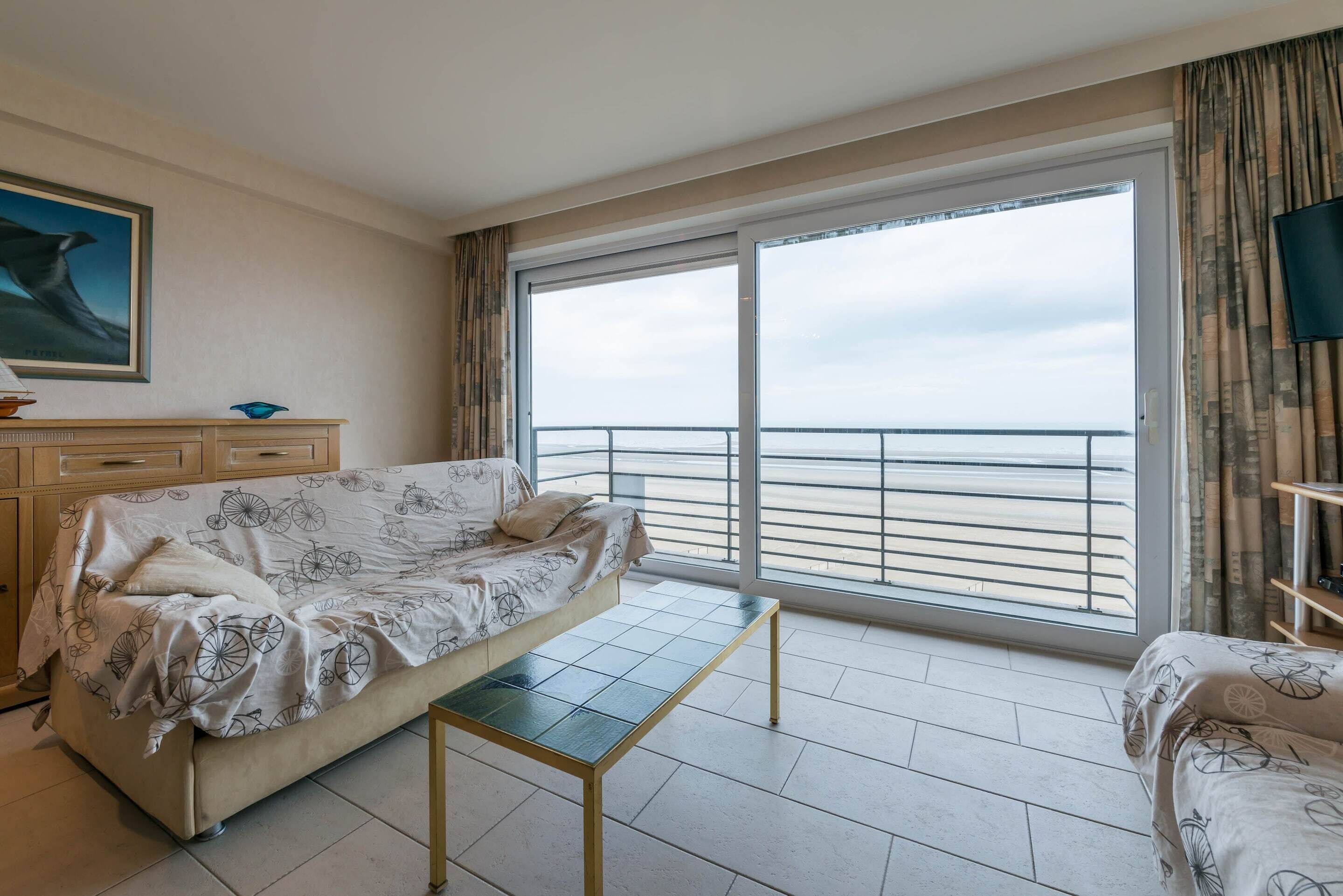 A hotel room with a bed, a small table, a dresser, and a large window with a view of the sea.