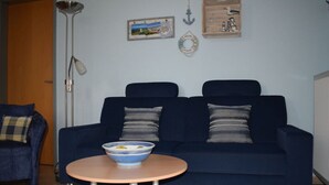 A living room with a dark blue sofa, a small round table with a bowl, a floor lamp, and wall decorations including a welcome sign and a framed picture. - Holiday Apartment Seagull (Zinnowitz)