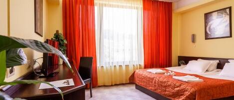 Habitación de hotel con una cama, un escritorio y una silla. Se ve una ventana con cortinas rojas, una planta y un cuadro en la pared.