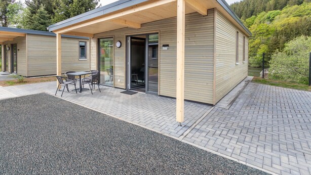 A modern building with a covered patio area, a small outdoor seating arrangement, and a paved walkway. - Familienfreundliche Bungalows am Kronenburger See (Hallschlag)