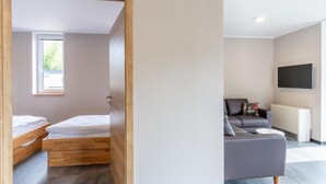 A modern, compact room with a wooden bed, a grey sofa, and a mounted TV. - Familienfreundliche Bungalows am Kronenburger See (Hallschlag)