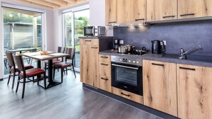 A modern kitchen with wooden cabinets, a built-in oven, and a dining area with a table and chairs. - Familienfreundliche Bungalows am Kronenburger See (Hallschlag)