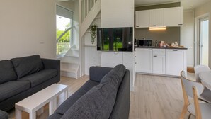 A modern living room with a grey sofa, a white coffee table, and a TV mounted on a white wall. A kitchen with white cabinets and a microwave is visible in the background.
