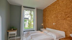 A bedroom with a bed, a bedside table with a lamp, a window with a view of greenery, and a wooden wall.