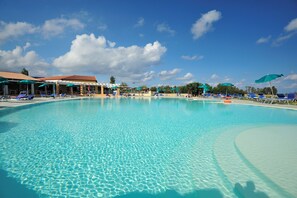 Una gran piscina con agua cristalina, rodeada de sillas reclinables y sombrillas azules, y un edificio con techo rojo al fondo.