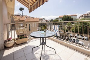 A balcony with a round glass table, potted plants, and a view of the street and buildings. - Homey Kiryat Haim Apt by Sea N Rent (Haifa)