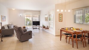 A spacious living area with a sofa, a dining table with chairs, a TV, and a balcony with a view. - Homey Kiryat Haim Apt by Sea N Rent (Haifa)