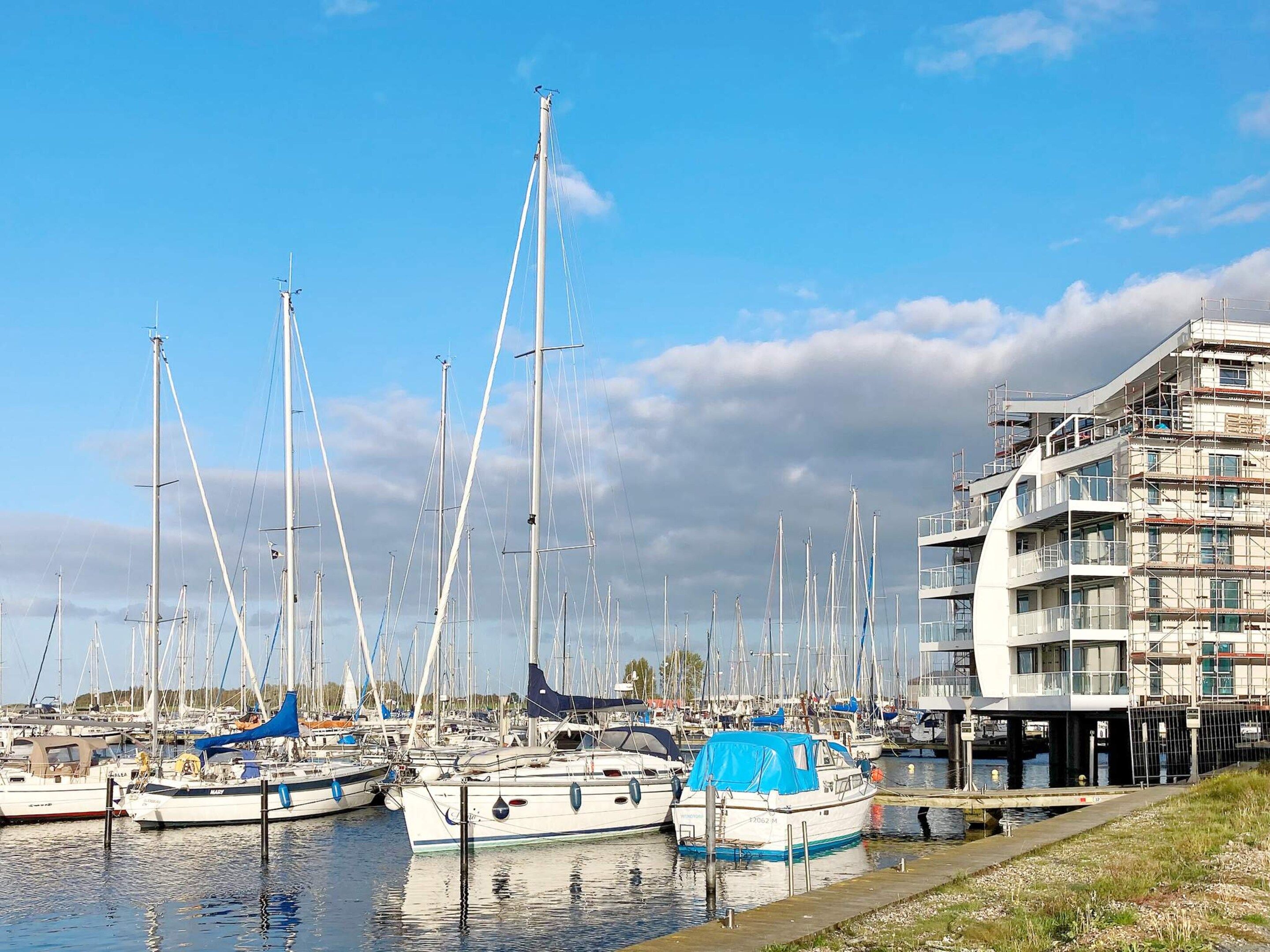 Een jachthaven met meerdere zeilboten afgemeerd, een modern gebouw met balkons en een heldere lucht.