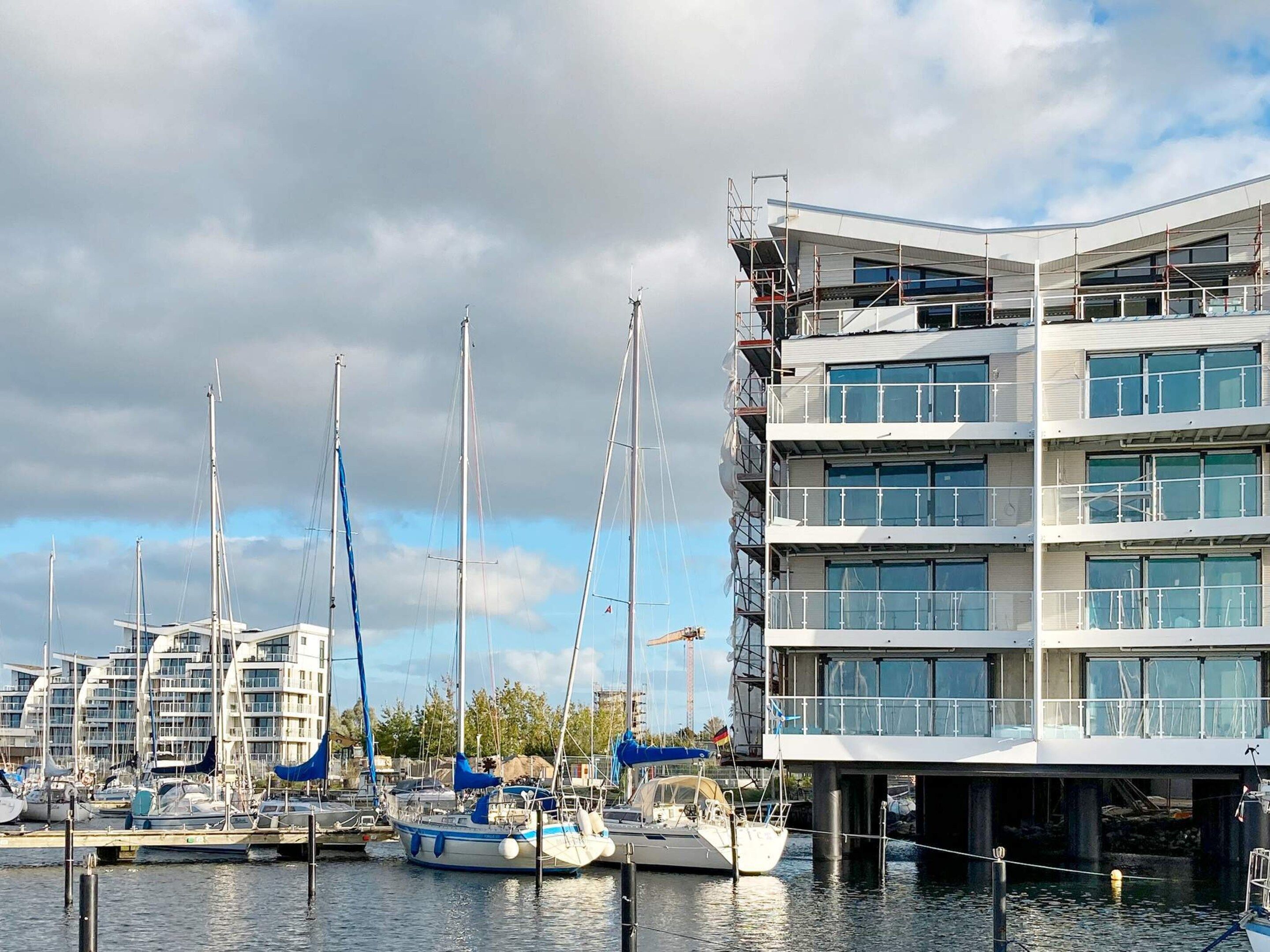 Un port de plaisance où sont amarrés de nombreux bateaux, un bâtiment moderne à étages et un ciel nuageux.