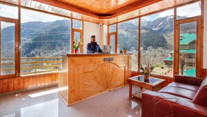 A reception area with a wooden reception desk, a sofa, and large windows offering mountain views. - La kailasha Regency (Manali)