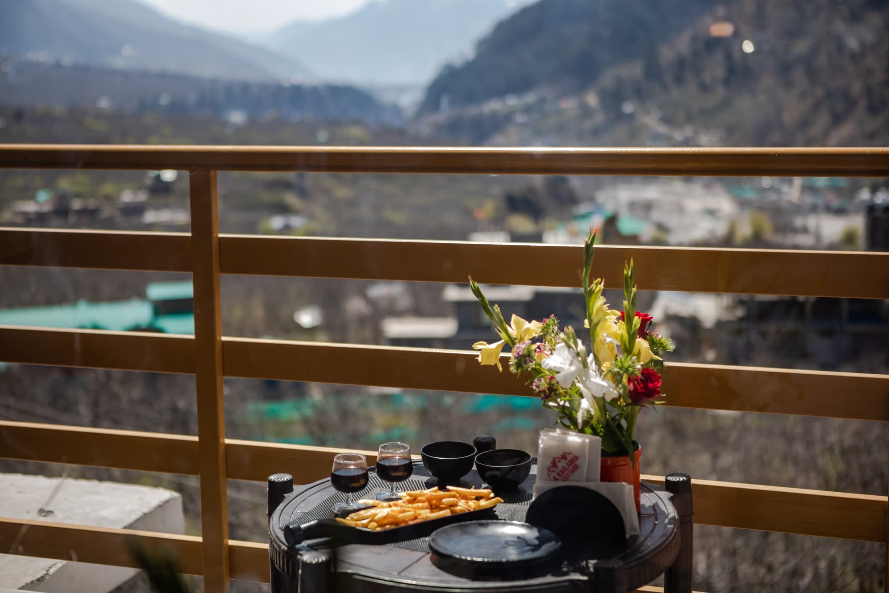 A table with a meal setup, including a plate of food, drinks, and a vase of flowers, viewed from a balcony with a scenic mountain backdrop.