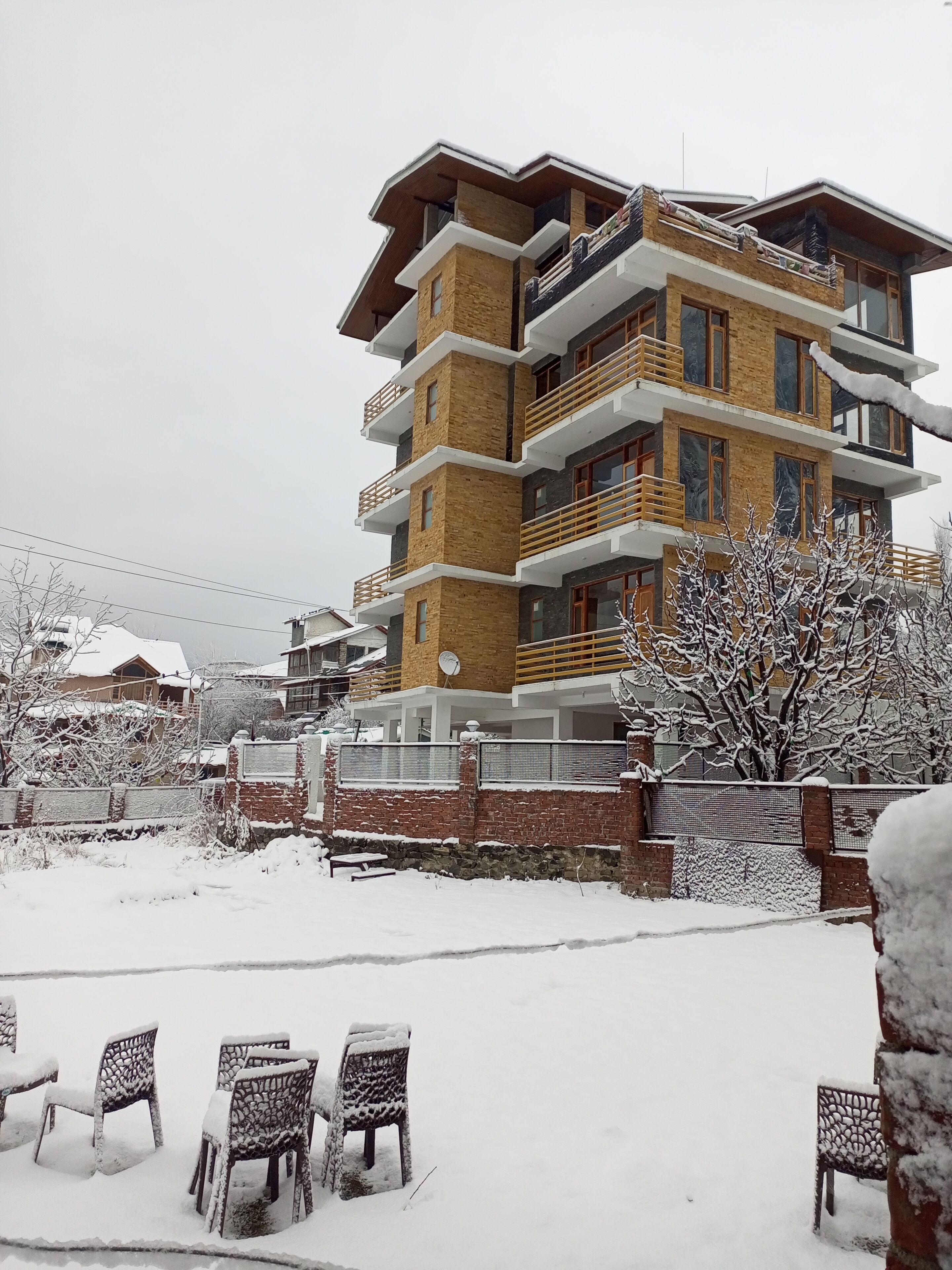 A multi-story residential building with balconies, a brick facade, and a snow-covered outdoor area with plastic chairs.