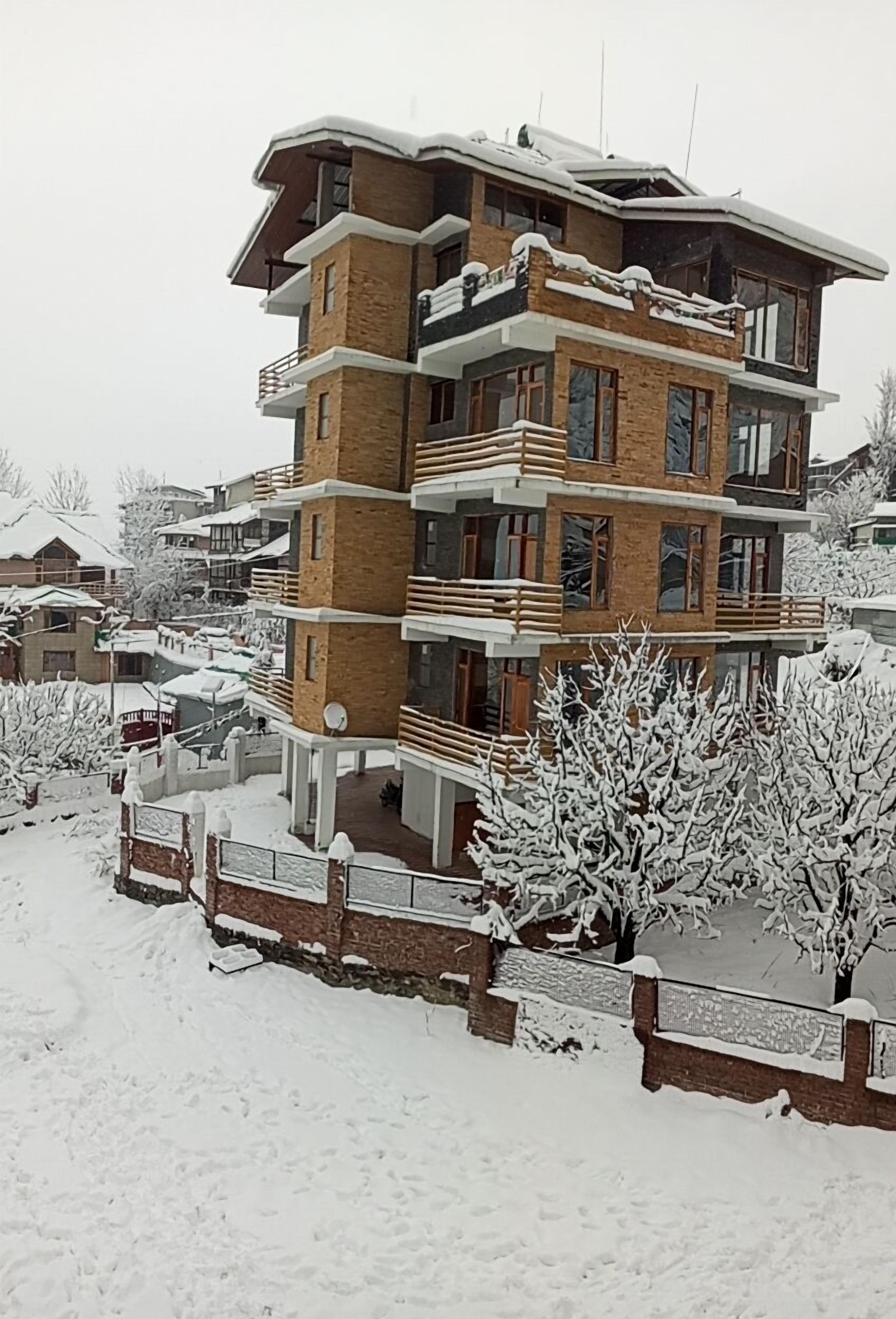 A multi-story building with a brick facade and balconies, surrounded by snow-covered trees and a fence.