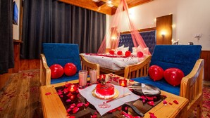 A room with a bed, two blue sofas, a wooden coffee table with a cake, candles, and red balloons. - La kailasha Regency (Manali)