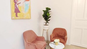Une pièce avec deux fauteuils roses, une petite table ronde sur laquelle se trouve un vase, une plante en pot et un tableau coloré accroché au mur.