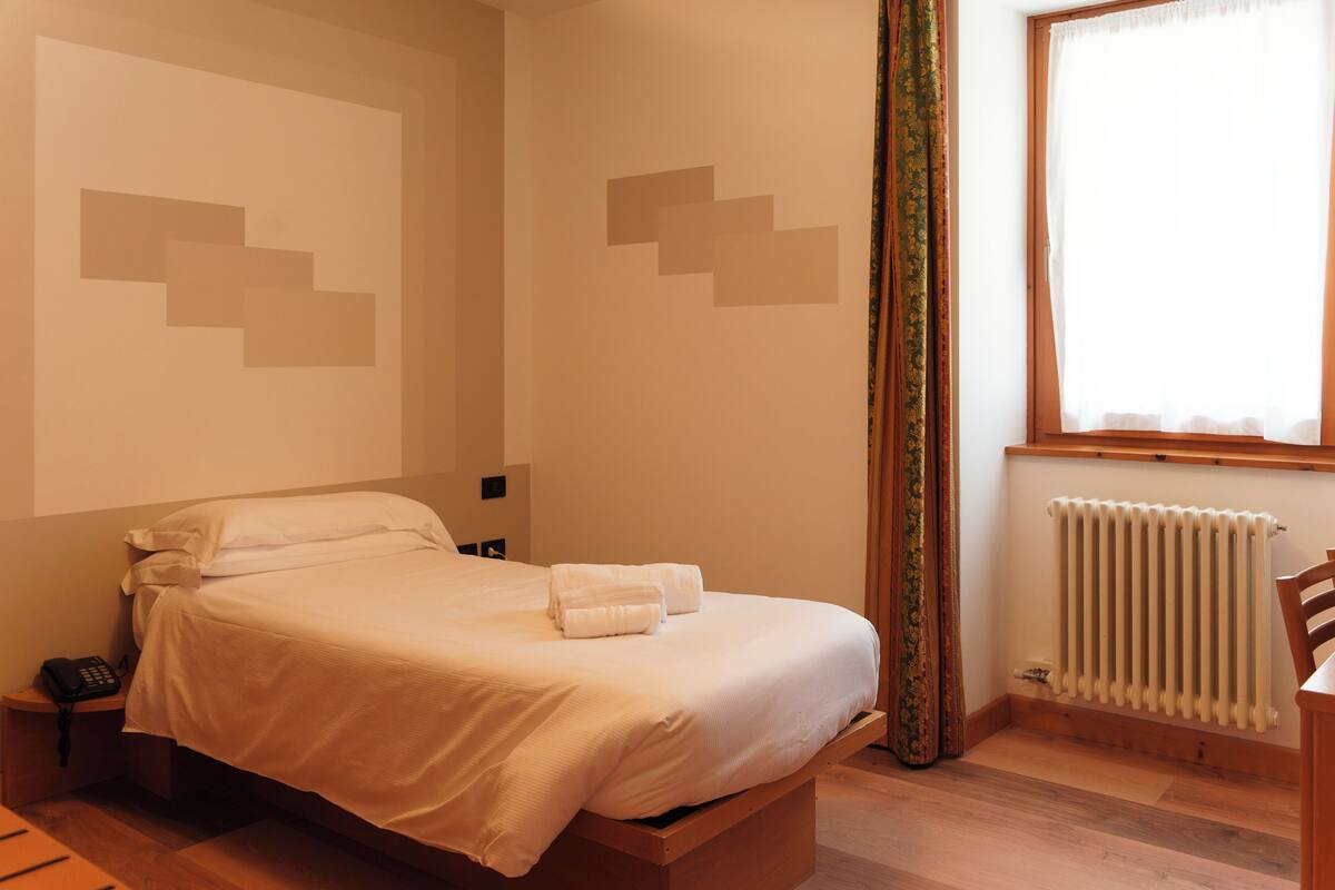 A neatly made bed with white linens and two folded towels, a bedside telephone, a wooden nightstand, a window with a wooden frame, and a radiator on the wall.