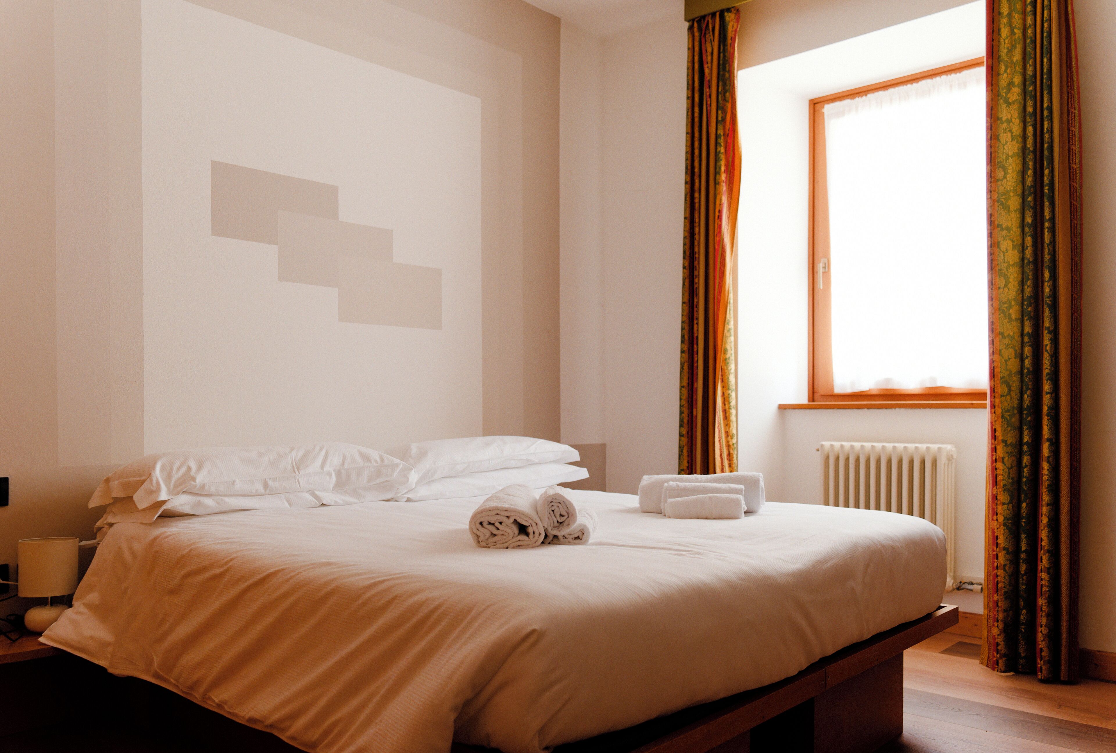 Una cama bien hecha con ropa de cama blanca y toallas enrolladas, una mesita de noche con una lámpara, y una ventana con cortinas.