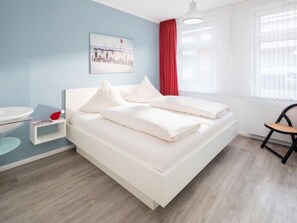A bedroom with a bed, a bedside table, a chair, a window with curtains, and a wall with a picture. - Frisian House Norderney (Norderney)