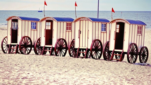 A row of beach cabanas with wheels, each with a door and windows, aligned on a sandy beach with the sea in the background. - Frisian House Norderney (Norderney)
