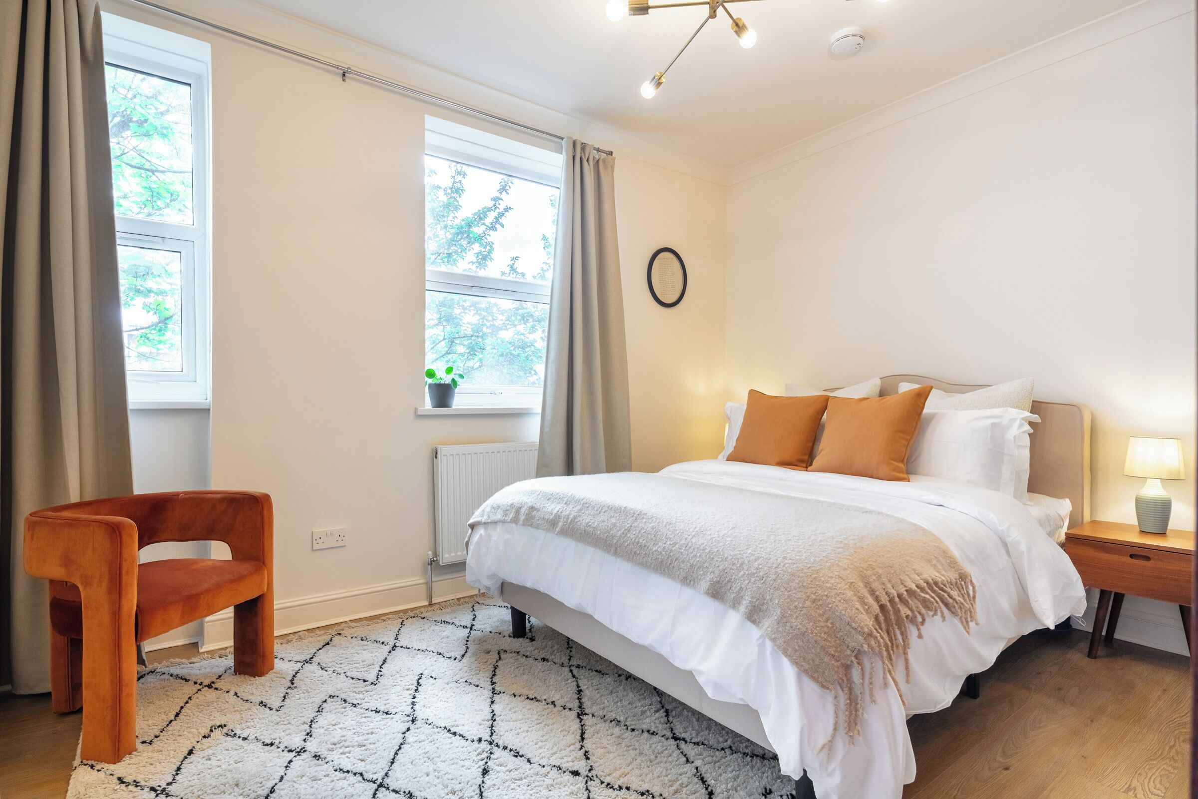 A bedroom with a bed, bedside table, lamp, chair, window, and a small plant.