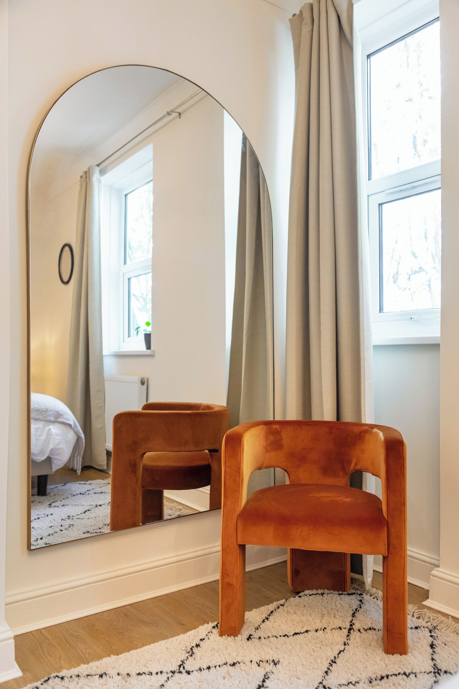 A room with a large mirror reflecting a bed and a window with curtains.